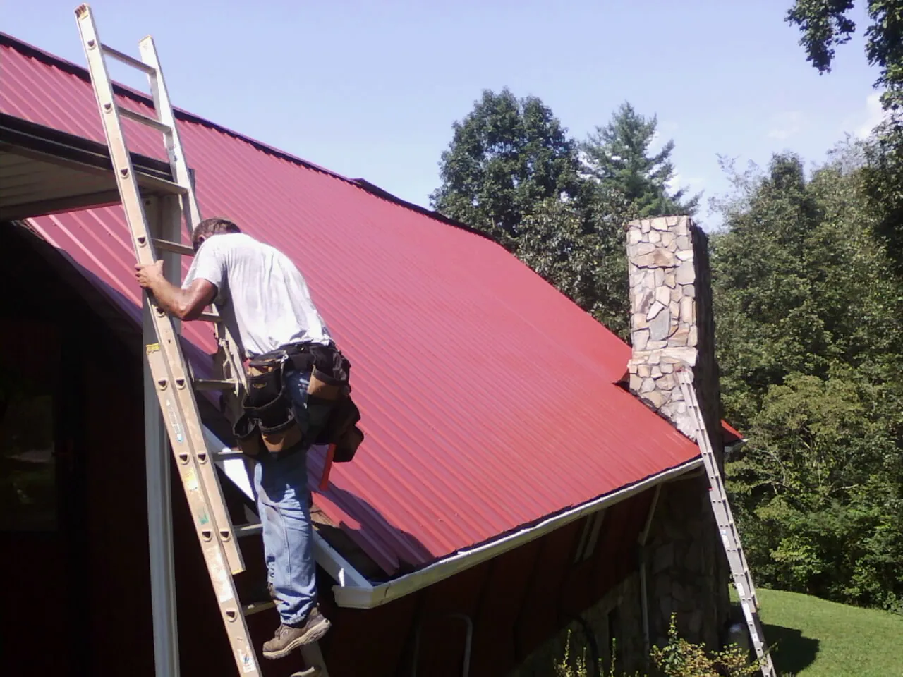 Roofer on a ladder beside a red metal roof and stone chimney — Soffit & Fascia Repair by Roofing Brothers in Burnet